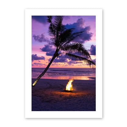 Photo of a fire pit by a palm tree on a beach with a sunset sky.